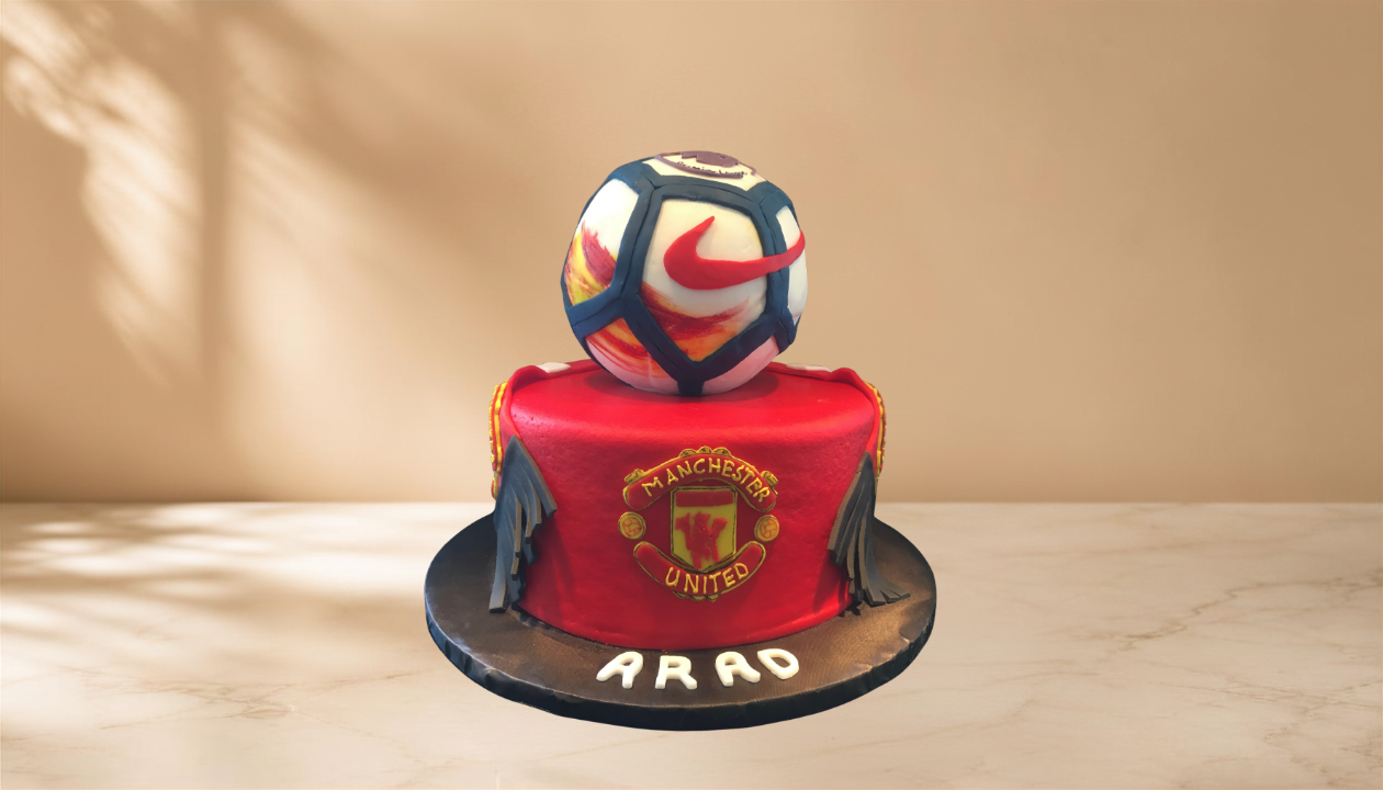 A cake decorated with the Manchester United football club logo, featuring a red and black color scheme, with a football on top and the name 'ARAD' written on the base.