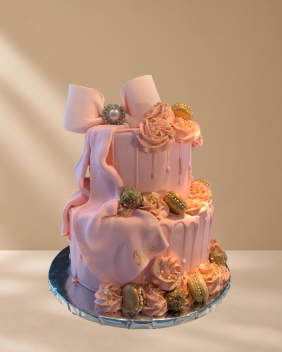 A two-tier pink cake decorated with pink frosting roses, macarons, and edible pearls, topped with a large pink bow.