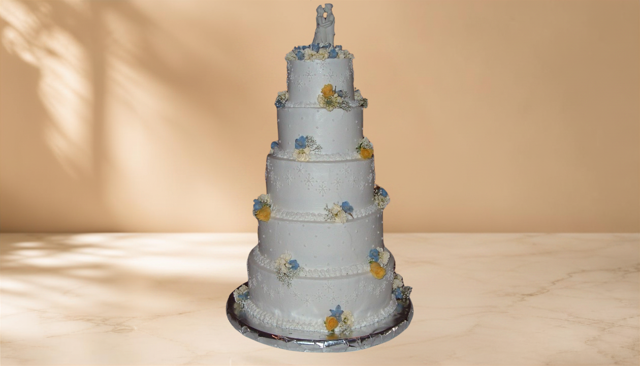 A tall, multi-tiered wedding cake with white icing, decorated with small yellow and blue flowers, and topped with a bride and groom figurine.