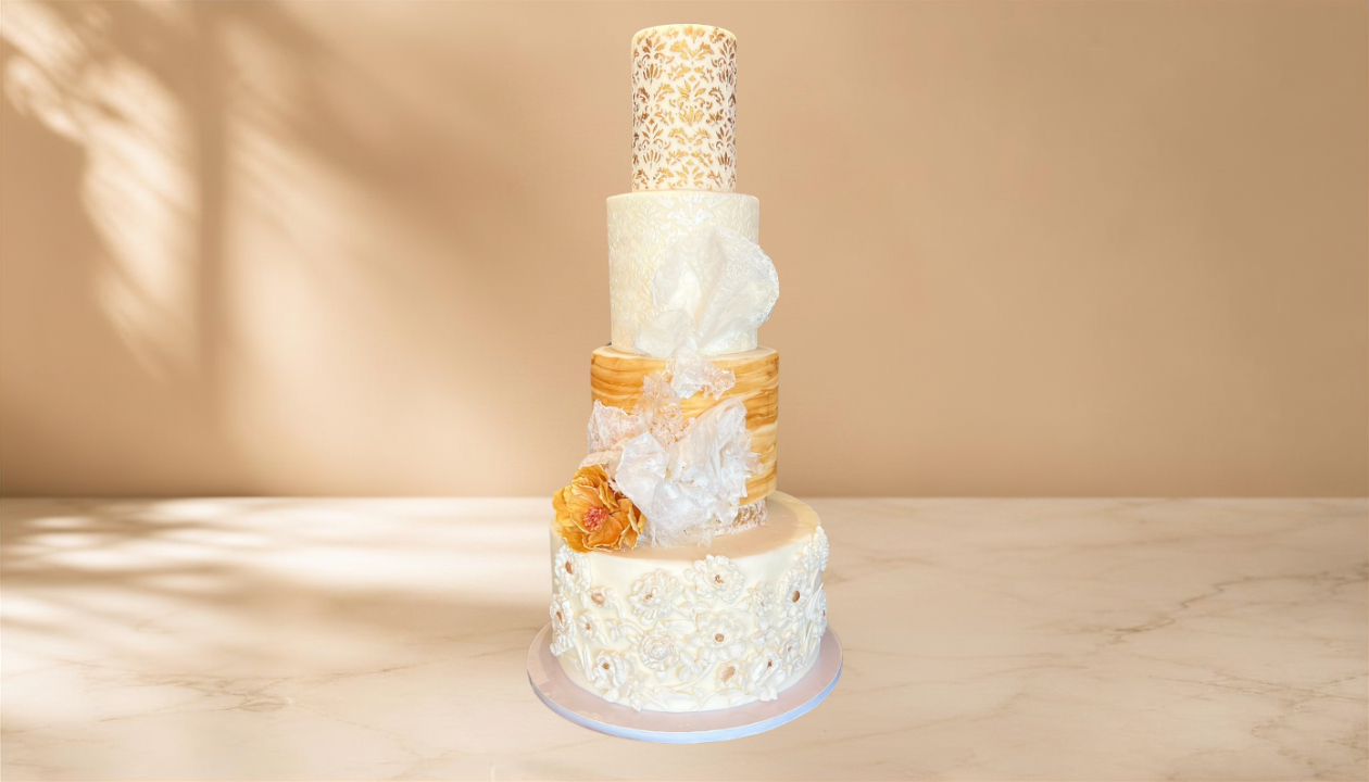 Elegant four-tier wedding cake with white and gold floral designs, decorative fabric, and sugar flowers, placed on a white marble surface with a beige background.