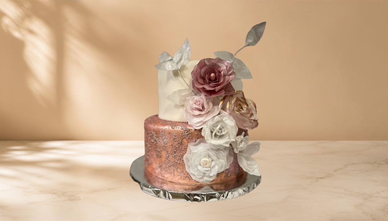A two-tiered cake with edible roses and leaves, featuring a gold accent and a marble design, displayed on a silver cake board on a light-colored table.