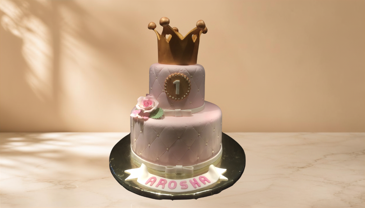 A two-tier pink cake with a gold crown on top, decorated for a first birthday celebration. The cake has a white border and a pink flower with green leaves on the side. A white fondant banner with the name "AROSHA" is at the base, and a round topper w