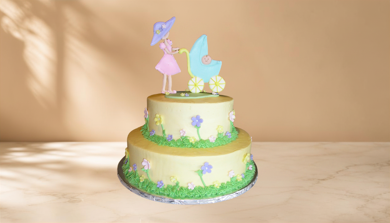 Yellow two-tiered cake decorated with pastel flowers and grass, topped with a figurine of a woman in pink holding a blue parasol and pushing a baby stroller.