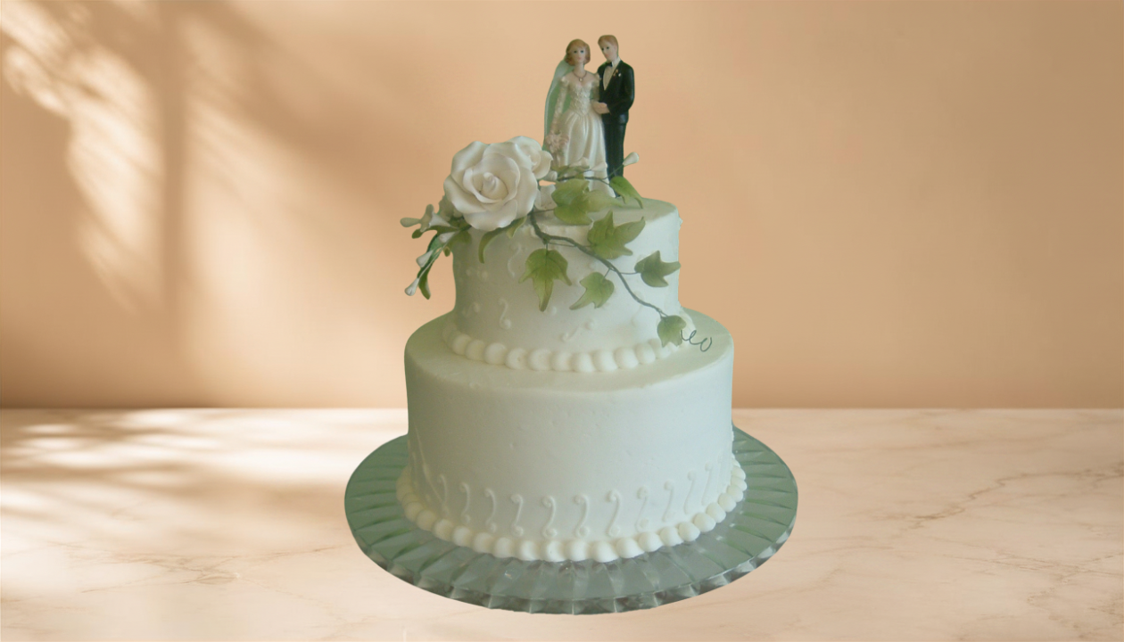 Two-tiered white wedding cake decorated with white roses and green leaves, topped with a figurine of a bride and groom.