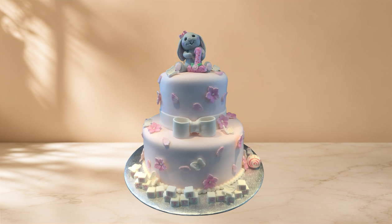 Two-tiered pastel-colored birthday cake decorated with pink flowers, a white bow, and a dog figurine holding a number one, set on a silver cake board on a marble surface with a beige background.