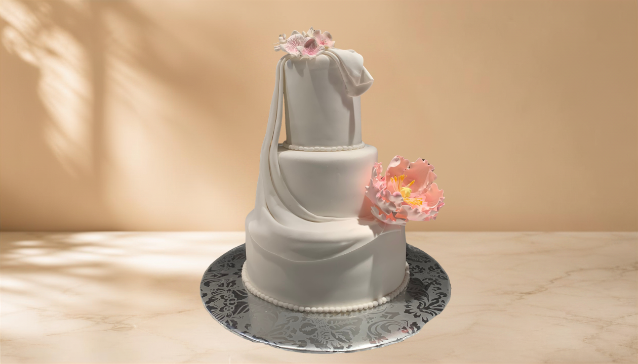 A white, multi-tiered wedding cake decorated with pink flowers and draped fondant, placed on a reflective silver cake board.