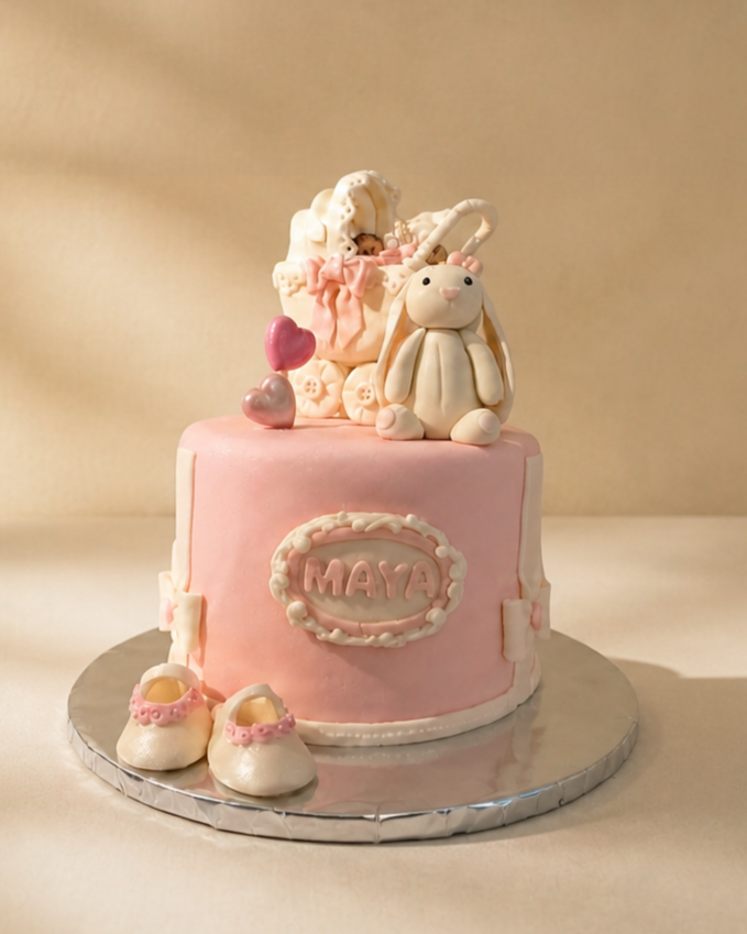 A pink baby shower cake with a baby bunny and baby bottle figurines on top, pink and silver heart decorations, a pair of baby shoes in front, and the name 'MAYA' on the front.