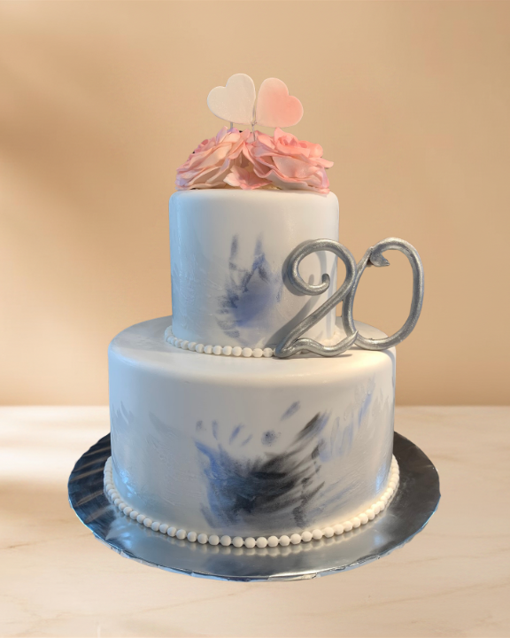 Two-tiered white birthday cake with blue and black marbling, pink icing flowers, heart and pink hearts on top, silver '20' cake topper, on a silver tray.