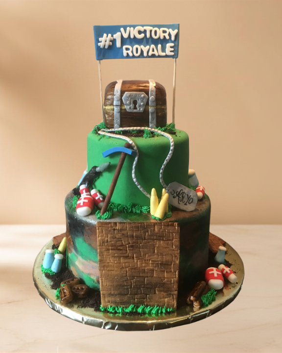 A two-tiered cake themed for the game Fortnite, decorated with gaming items, a treasure chest on top with a sign that says '#1 Victory Royale', and various elements related to the game including pickaxes, shoes, and a shield.