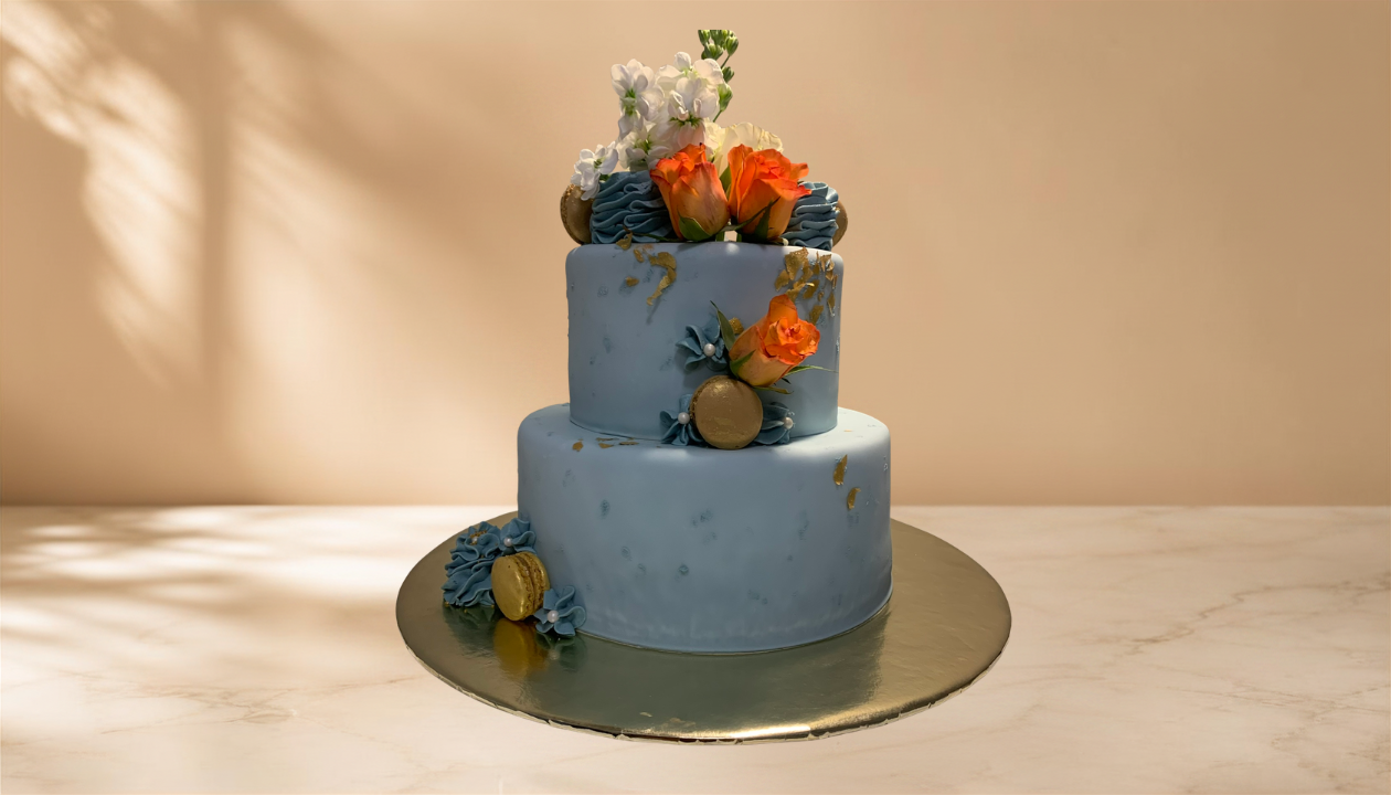 Two-tiered blue cake decorated with orange roses, white flowers, gold accents, and blue frosting details, set on a silver cake board.