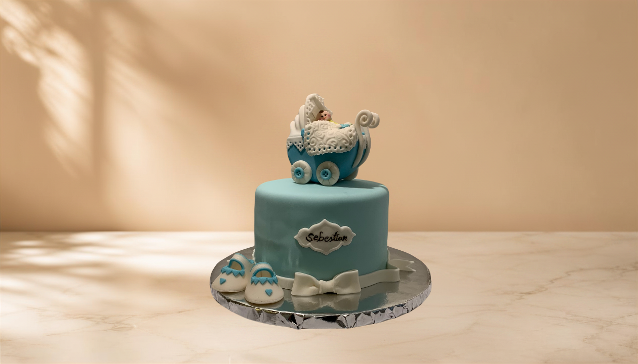 A light blue cake with a toy baby stroller on top, decorated with white and blue fondant, placed on a reflective silver cake board. The cake has a white bow at the base and a small, decorative plaque with the name "Sebastian" written on it, alongside