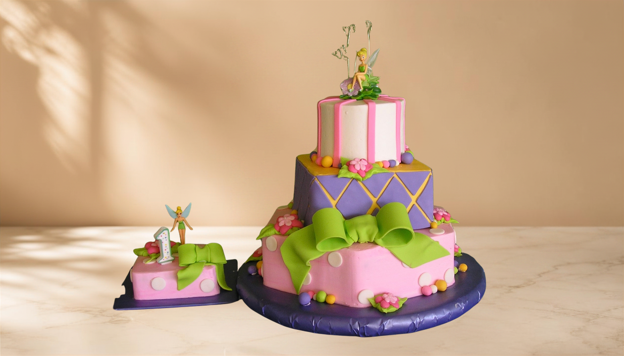 Colorful fairy-themed birthday cake with three tiers, decorated with pink, purple, and green fondant, featuring fairy figurines, floral decorations, and a large green bow, on a round blue cake base.