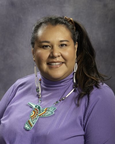 Portrait of a woman with long dark hair in a ponytail, wearing a purple turtleneck and a colorful hummingbird necklace, smiling against a gray background.