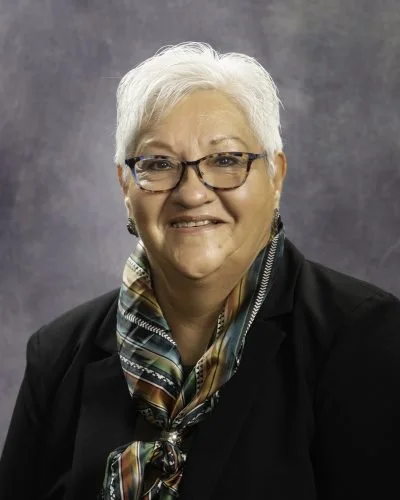 Portrait of an older woman with white hair, glasses, wearing a black jacket and colorful scarf, smiling against a gray background.