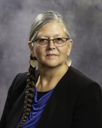 Portrait of a middle-aged woman with glasses and long braided hair, wearing a black blazer and a blue top, against a dark gray background.