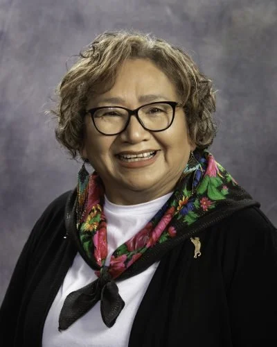 A smiling woman with curly hair, glasses, wearing a colorful scarf and a black jacket, against a gray background.