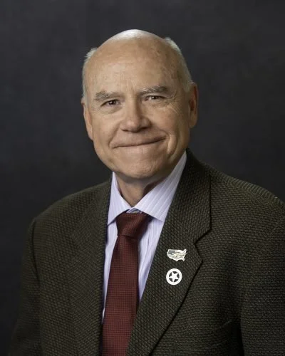 An elderly man with a bald head, wearing a dark suit jacket, a light blue shirt, and a maroon tie, posing against a dark background.