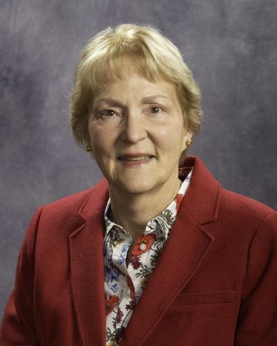 A portrait of an older woman with short blond hair, wearing a red blazer and a floral shirt, against a gray background.