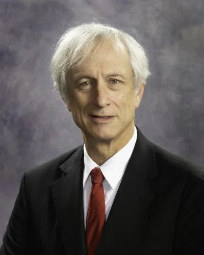 A man in a black suit, white shirt, and red tie with gray hair, standing against a gray background.