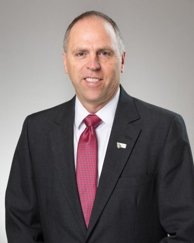 Professional man in a dark suit, white shirt, and red tie, smiling, with a gray background.