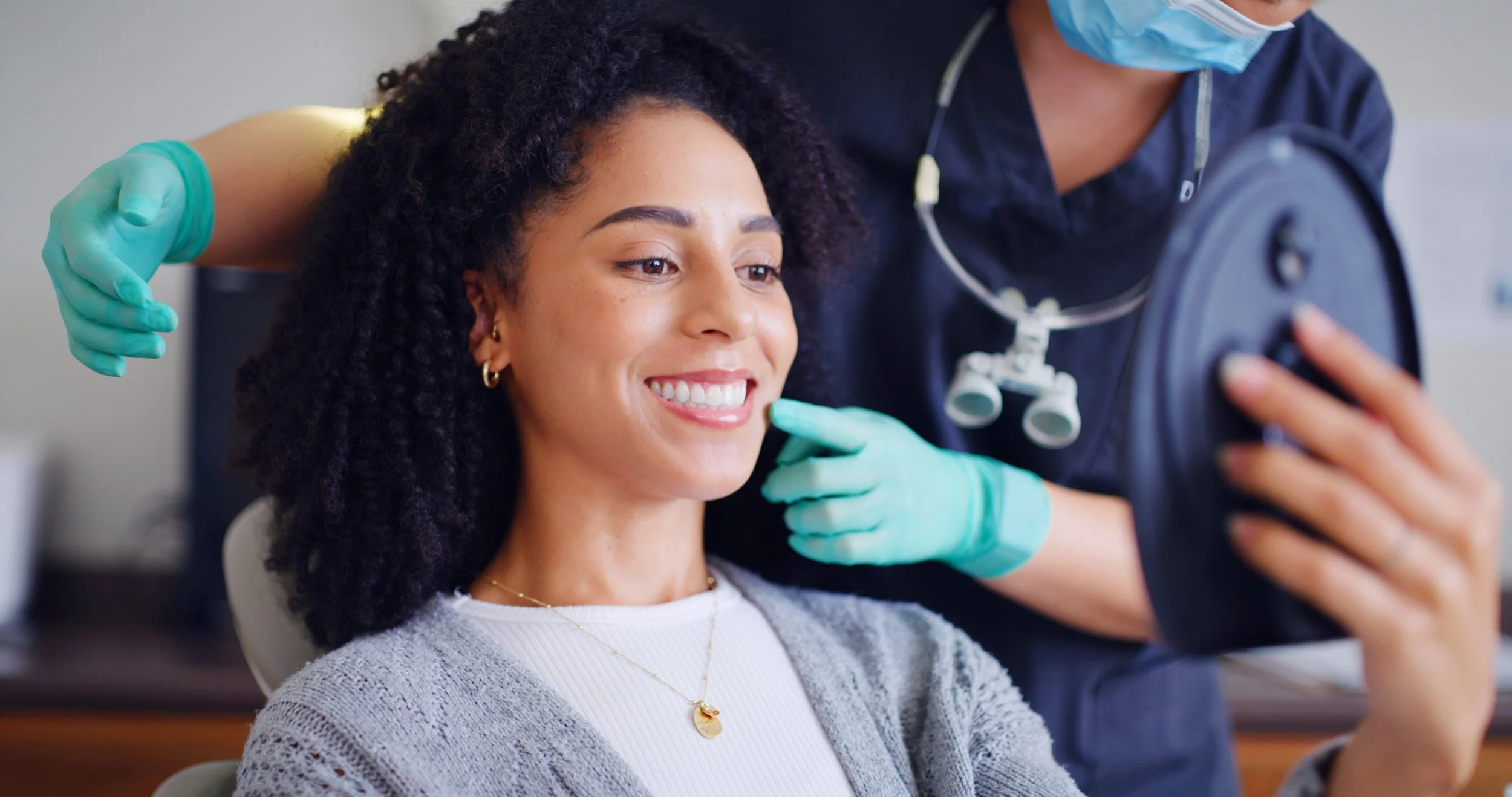 A woman sitting in a dental office smiling while a dental professional examines her teeth. The dental professional is wearing gloves and a mask, and is holding dental tools.