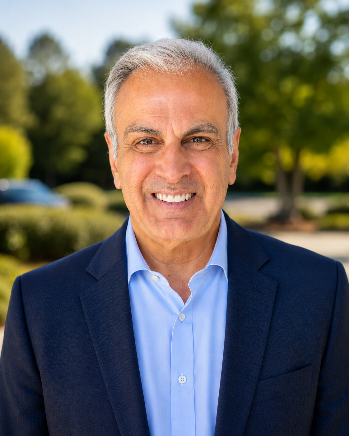 A middle-aged man with gray hair wearing a dark suit and light blue shirt smiling outdoors with trees and a blurred car in the background.