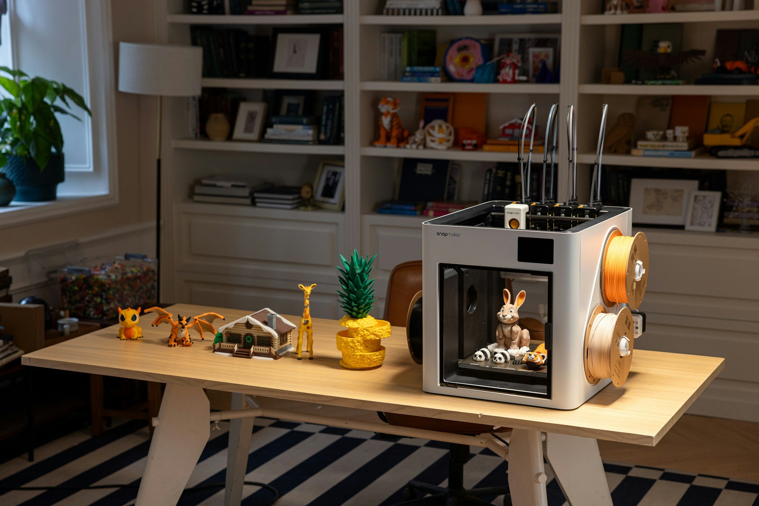A wooden table with small toy animals and buildings, along with a 3D printer holding spools of filament in a room with bookshelves, a window, and a striped black and white rug.