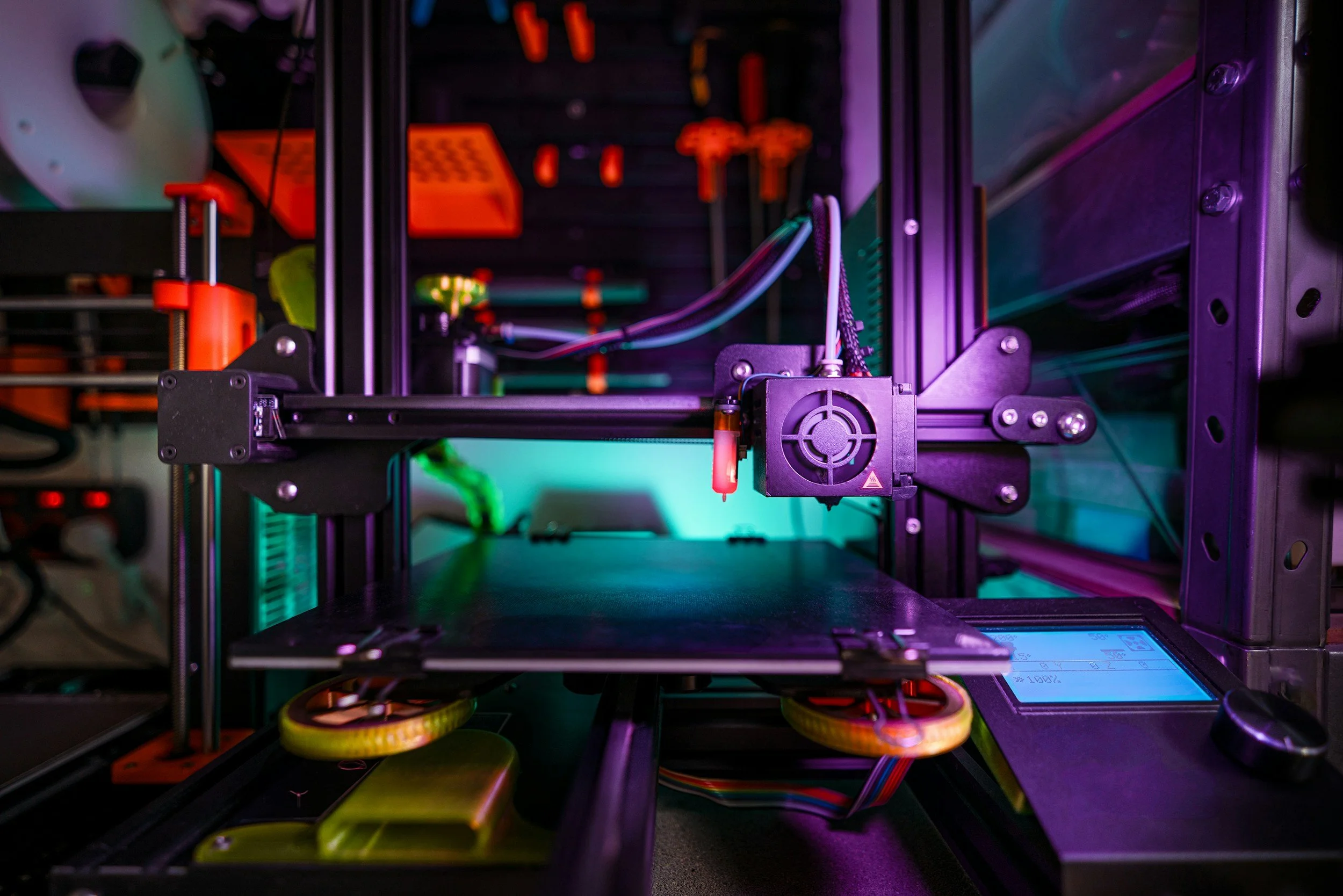 Close-up of a 3D printer's internal components with colorful wiring, orange parts, and a digital display, illuminated by multicolored LED lights.