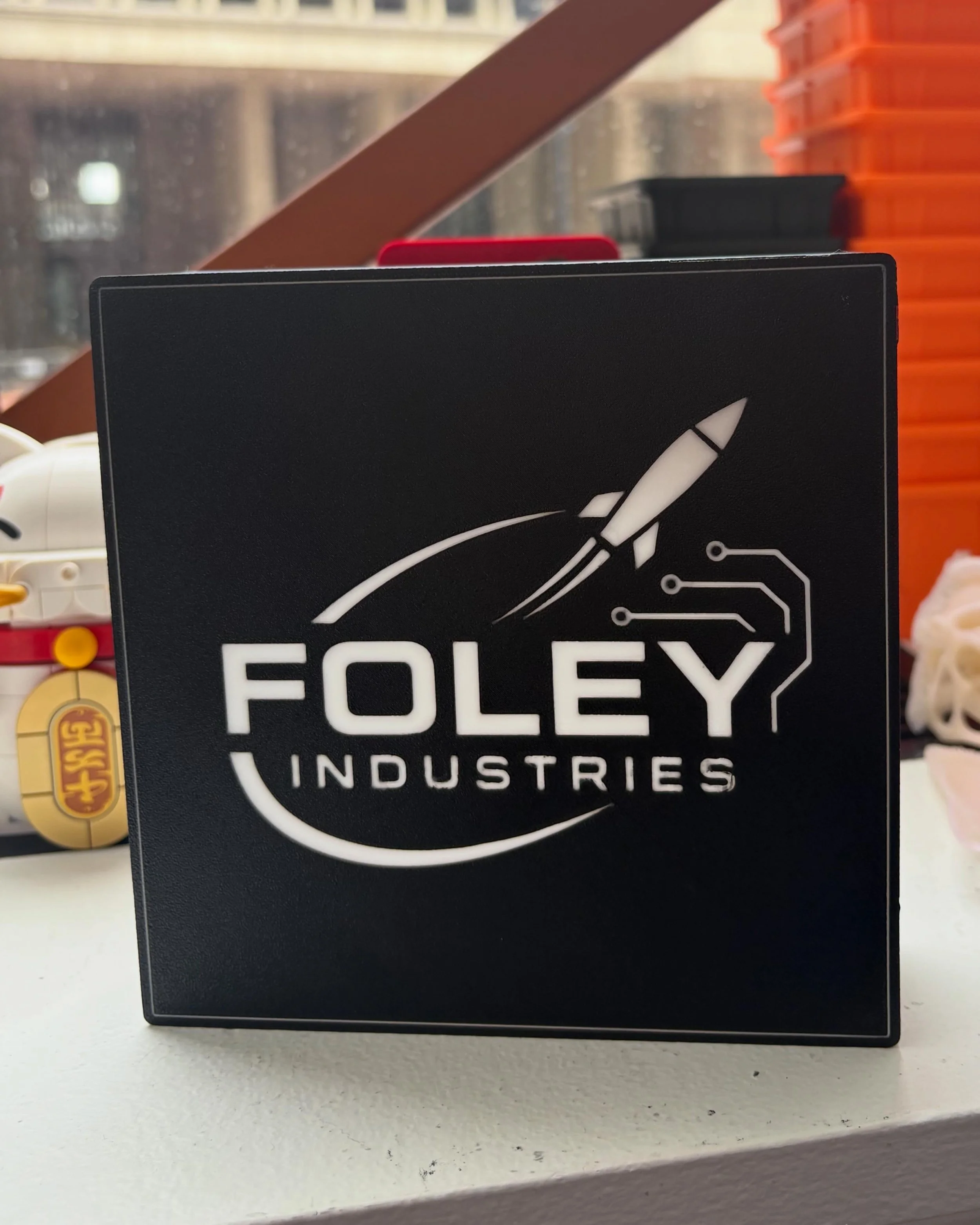 Close-up of a black rectangular object with a white rocket icon and the words 'FOLXY Industries' on it, placed on a white surface. In the background, part of a LEGO No. 3-15-2 set and some orange storage containers are visible.