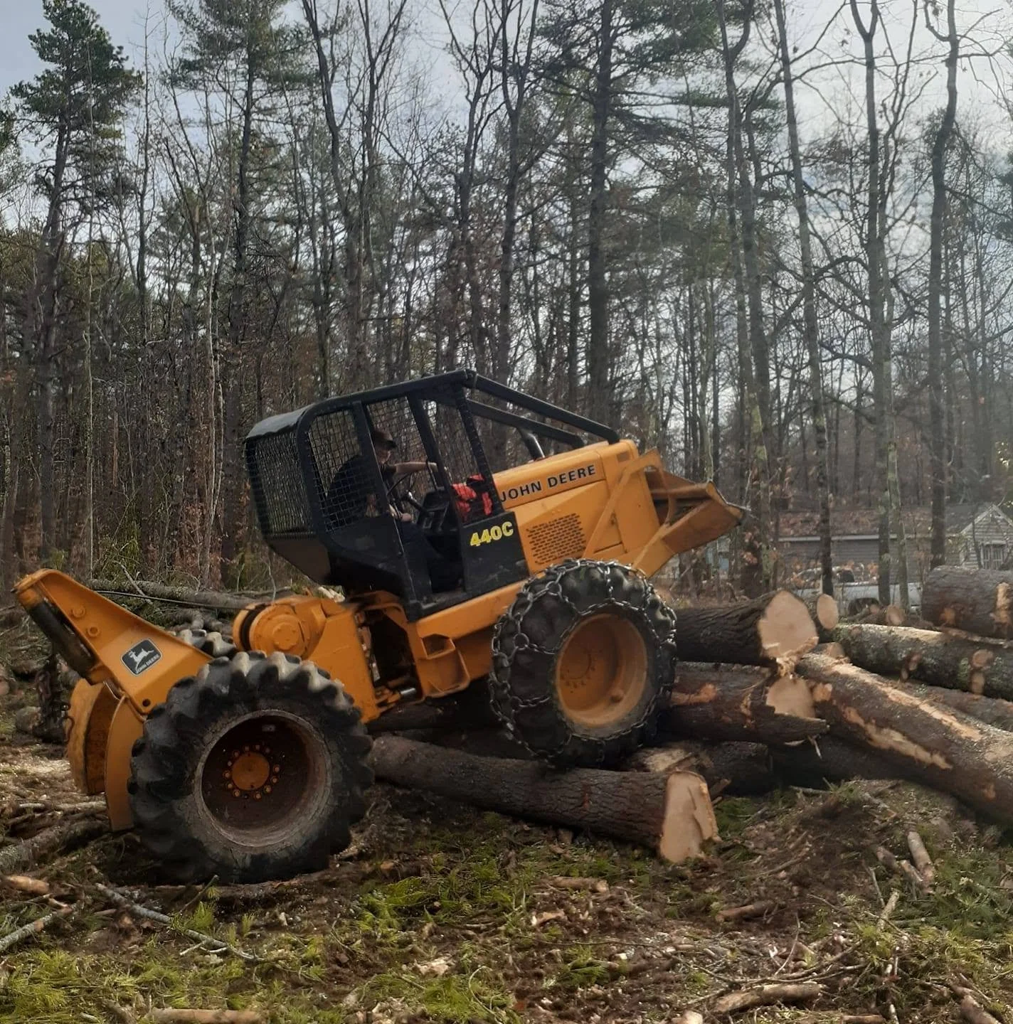 Yellow John Deere 440C skid steer loader clearing fallen trees and debris in a wooded area.