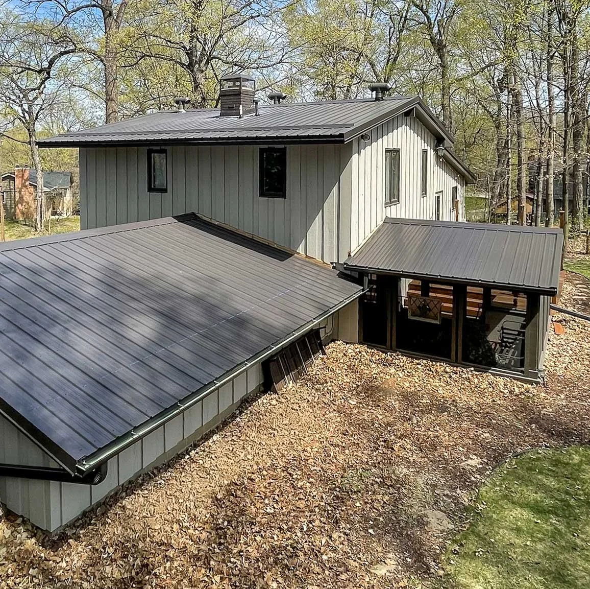 A two-story house with metal roofing, surrounded by trees and a backyard area covered with fallen leaves, in a suburban setting.
