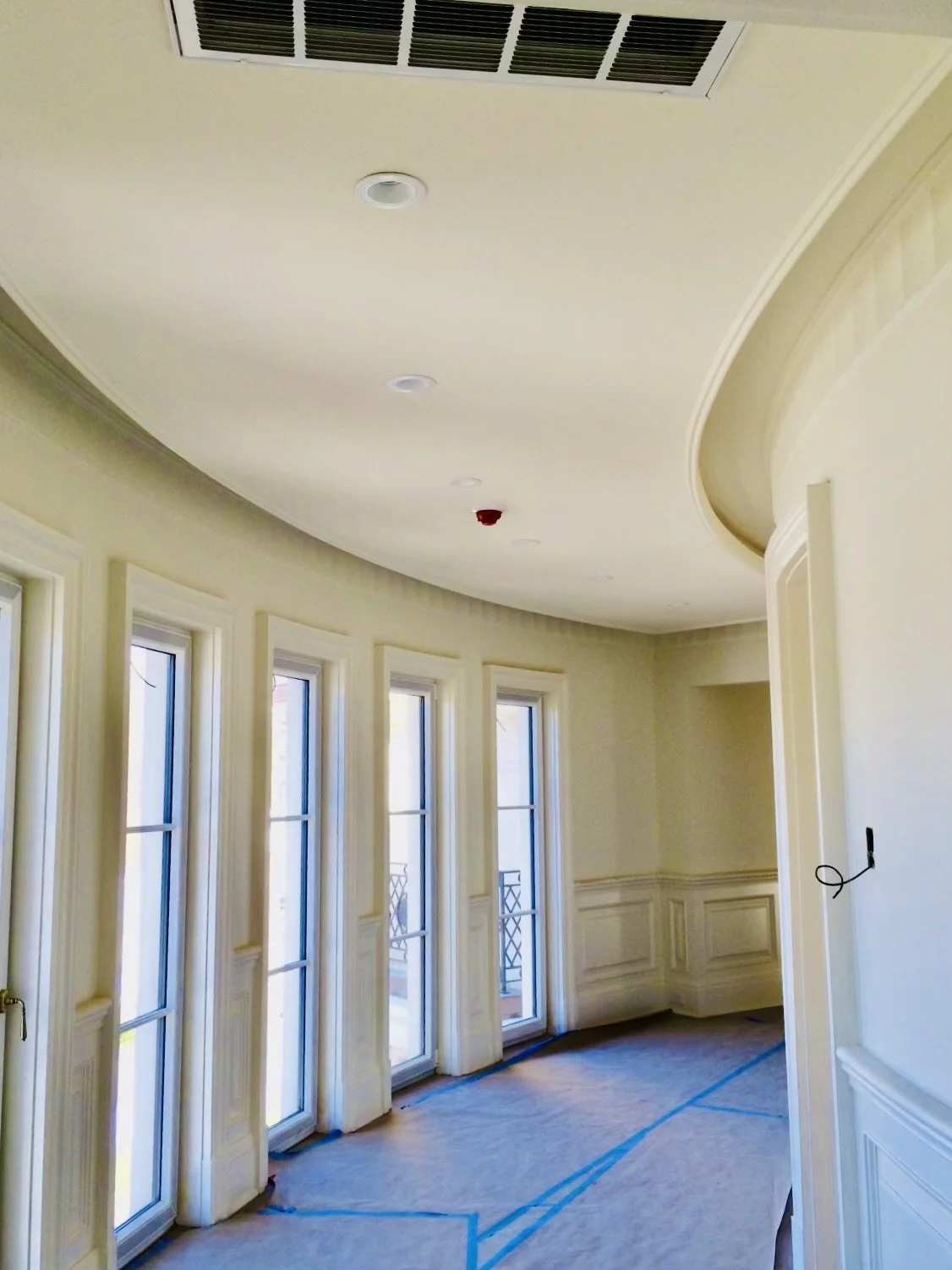 Empty room with tall windows, crown molding, an air vent on the ceiling, and a blue tape outline on the floor, possibly under construction or renovation.