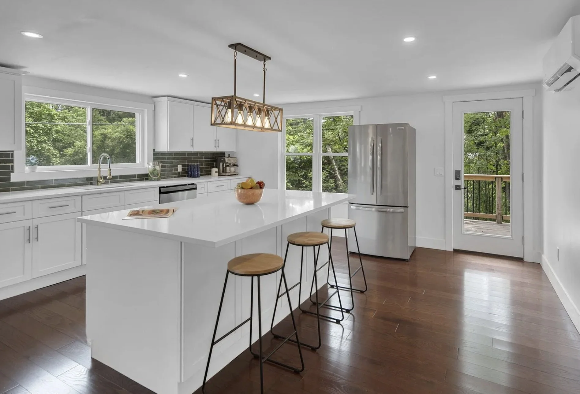 Modern kitchen with white cabinets, a white island with a bowl of fruit, stainless steel refrigerator, large windows showing green trees, wooden flooring, and a door leading outside.