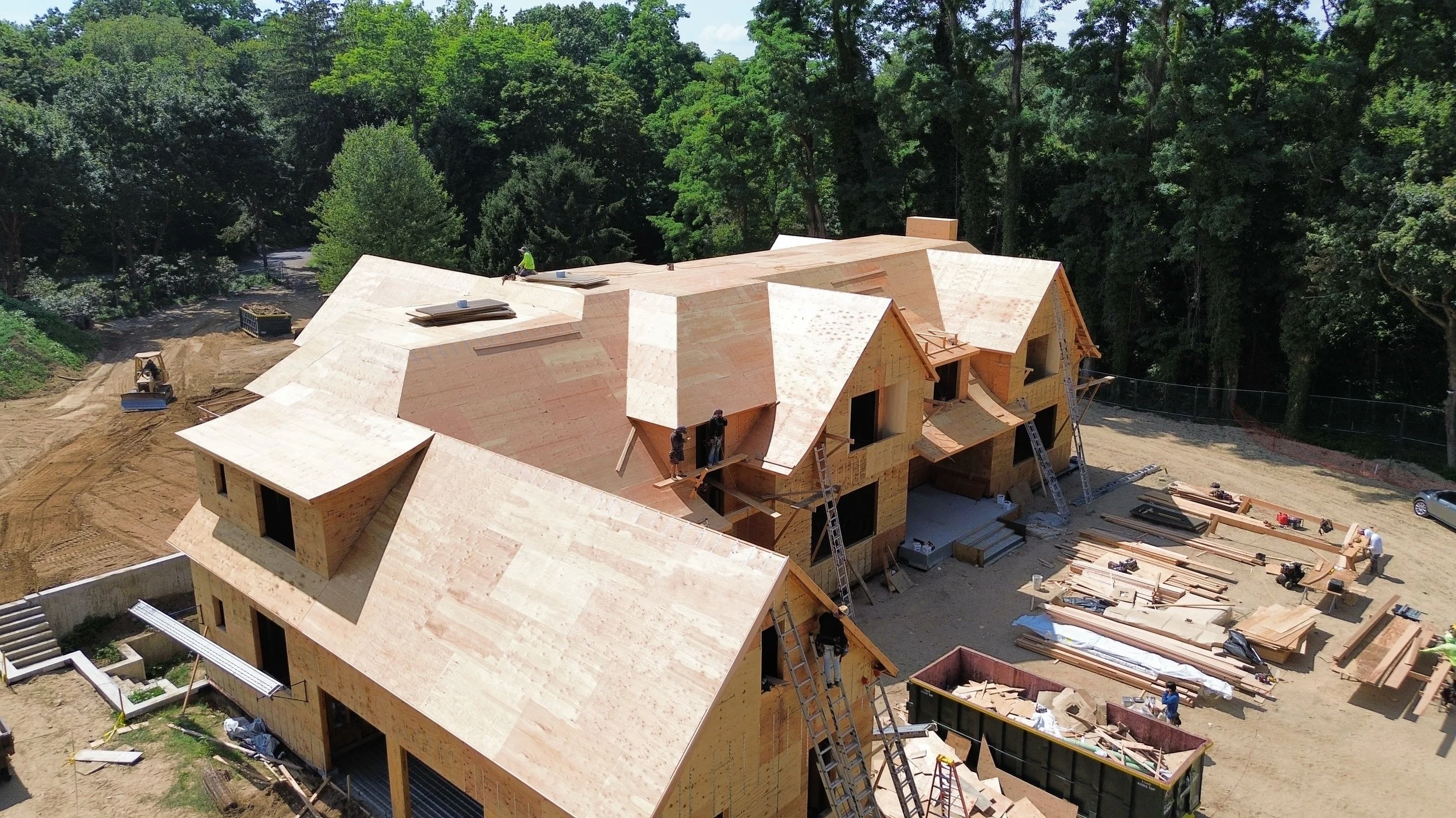 A large house under construction with a complex roof structure, surrounded by a forested area.