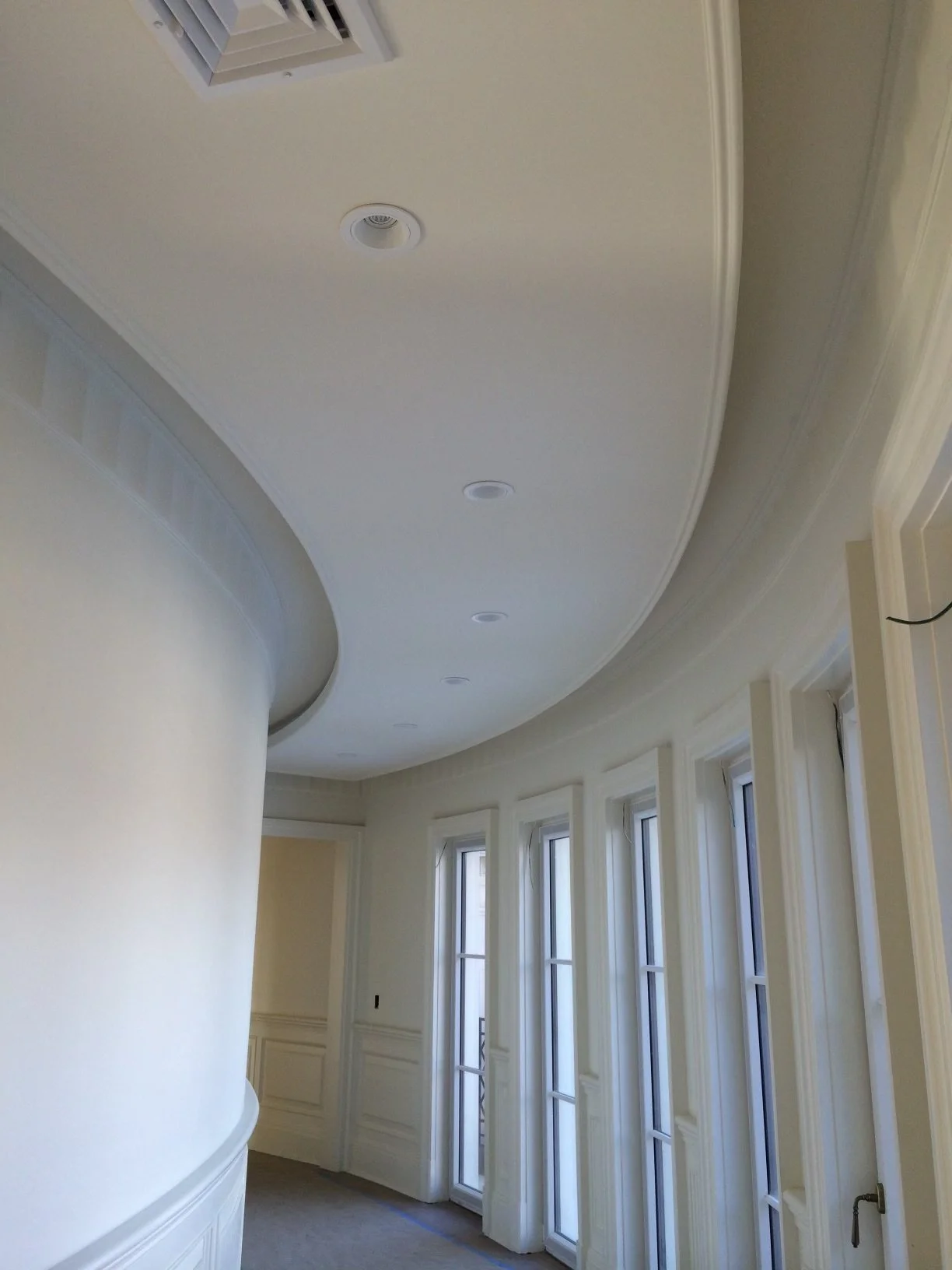 Interior view of a room with a curved ceiling, tall windows with white trim, and multiple ceiling lights. The walls are painted in a light color, and there is crown molding along the ceiling. The room appears to be under construction or renovation.