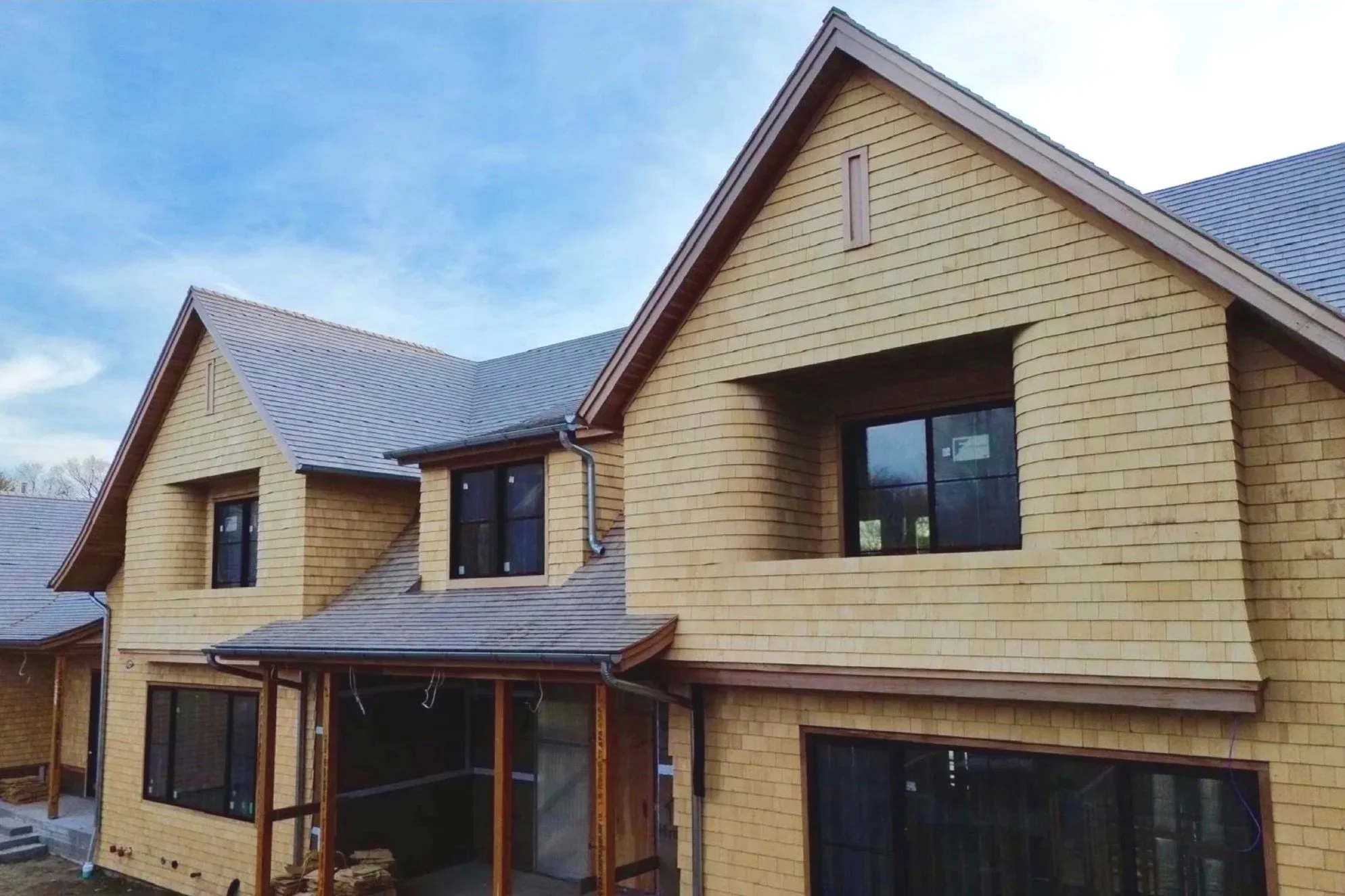 A multi-story house under construction with yellow brick exterior and dark gray roof shingles, featuring multiple windows and a covered patio area.