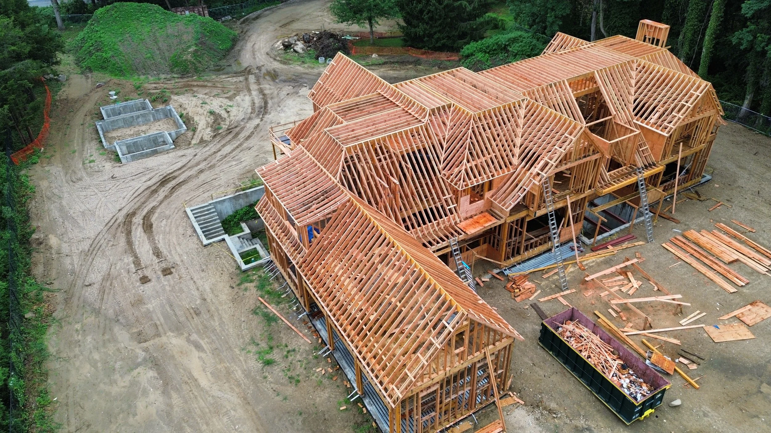 Aerial view of a house under construction with wooden framing and roof trusses, surrounded by dirt and construction materials.