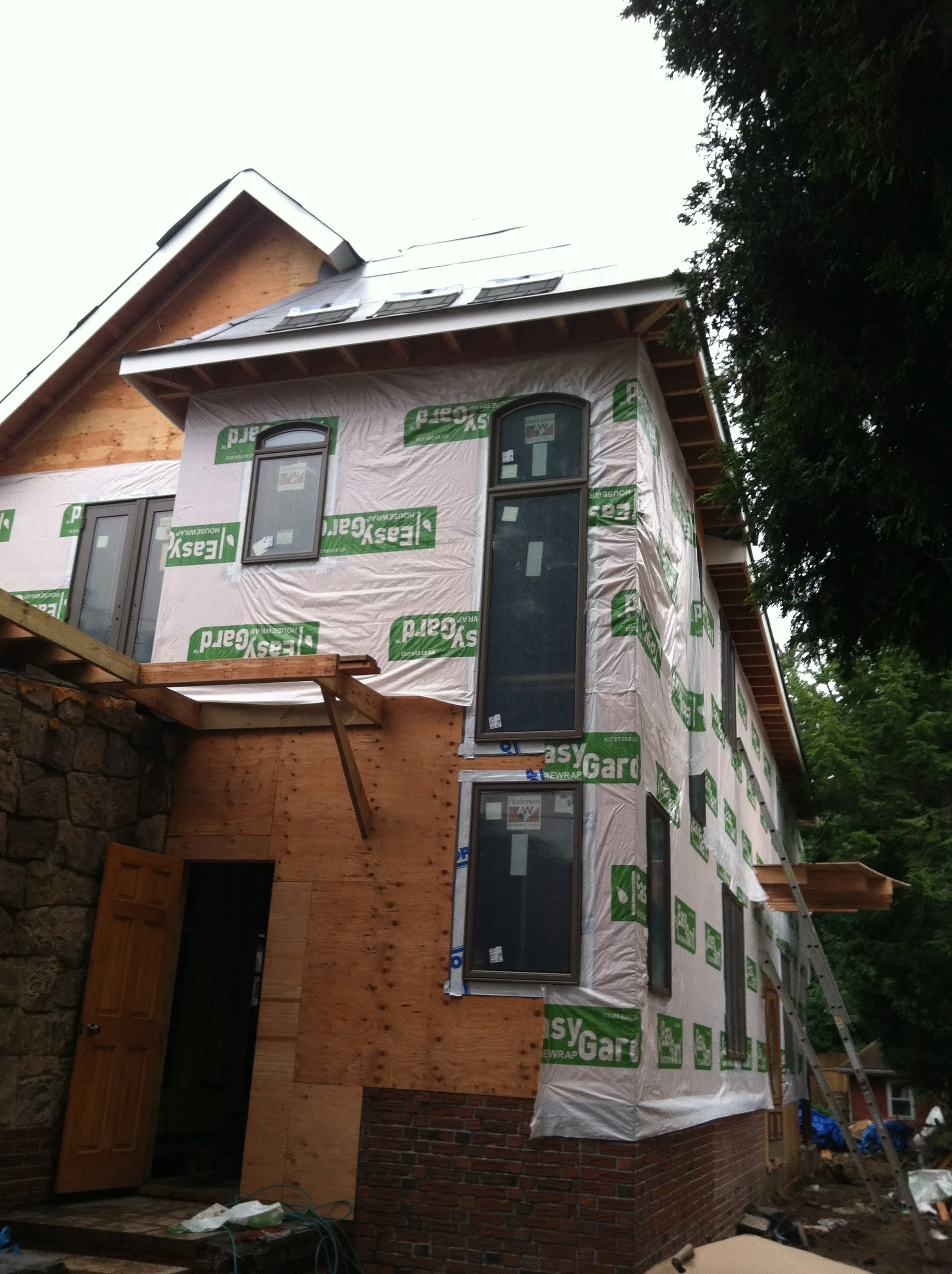 A house under construction with partially finished brick and wooden siding, with window frames installed and a ladder leaning against the building.