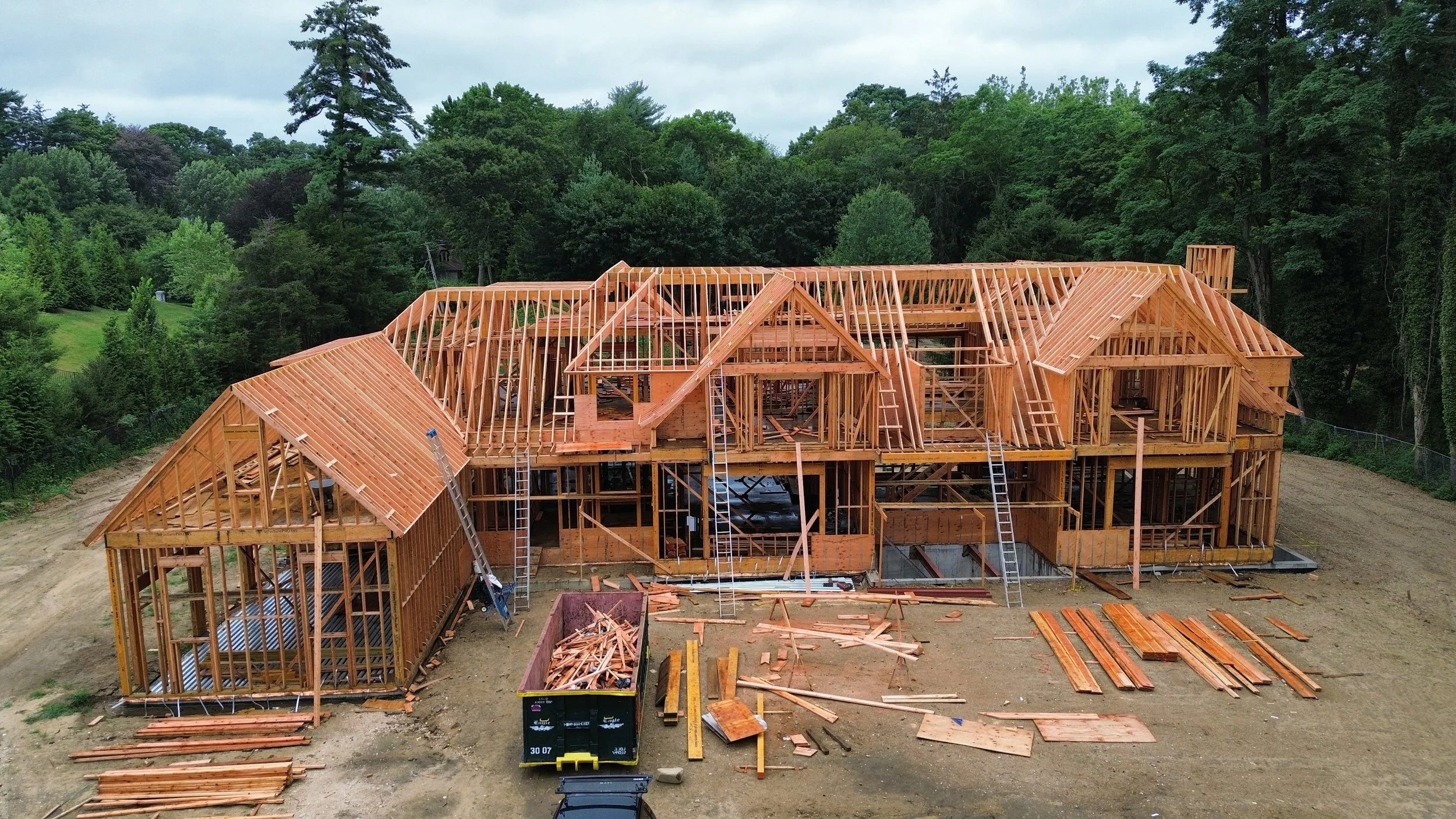 A large wooden house under construction in a wooded area with multiple levels, construction materials, and ladders visible.