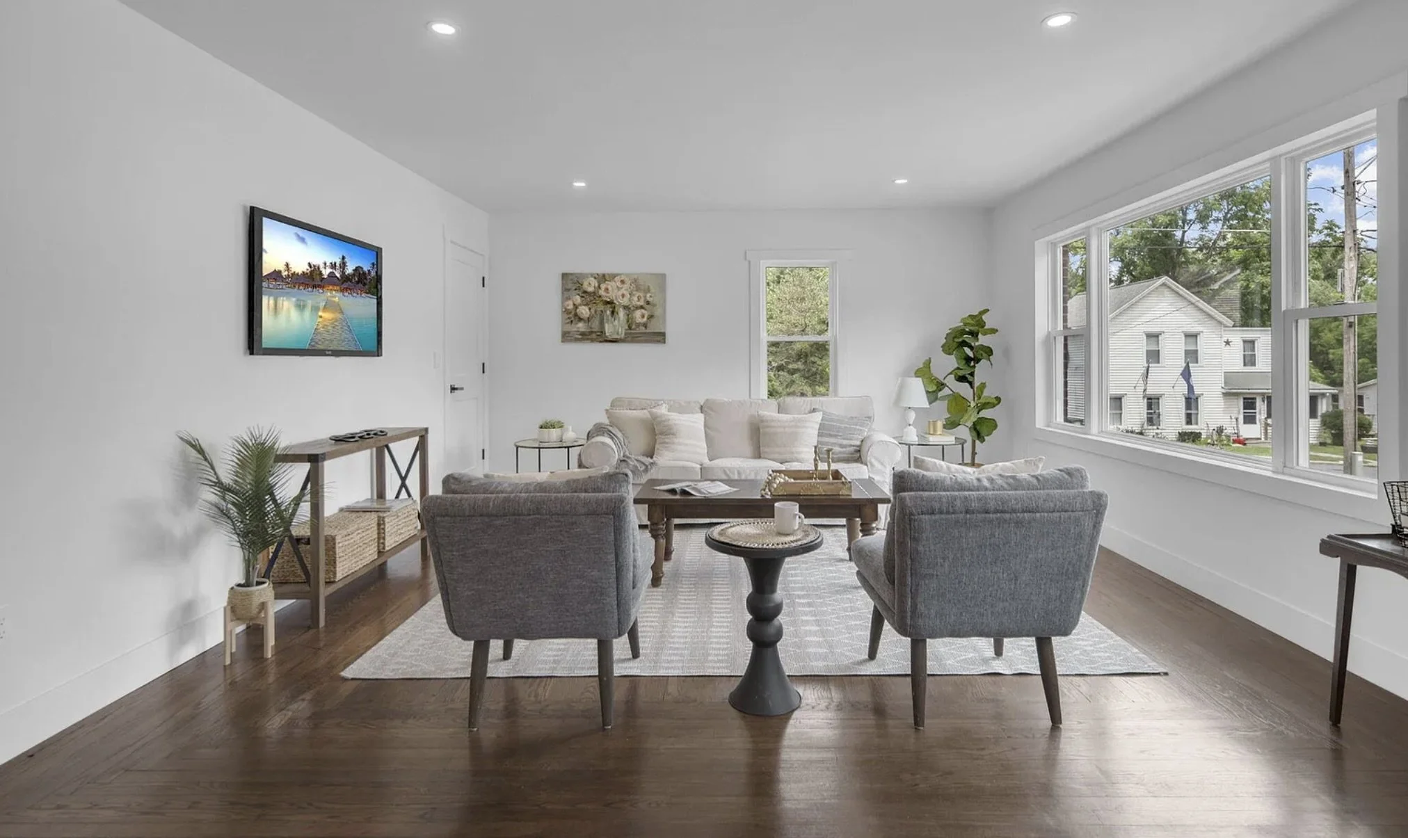Living room with white walls, hardwood floors, a beige sofa with pillows, four gray armchairs, a round black side table, a rectangular coffee table, a TV mounted on the wall, window with view of neighboring houses and trees, potted plant, and wall ar