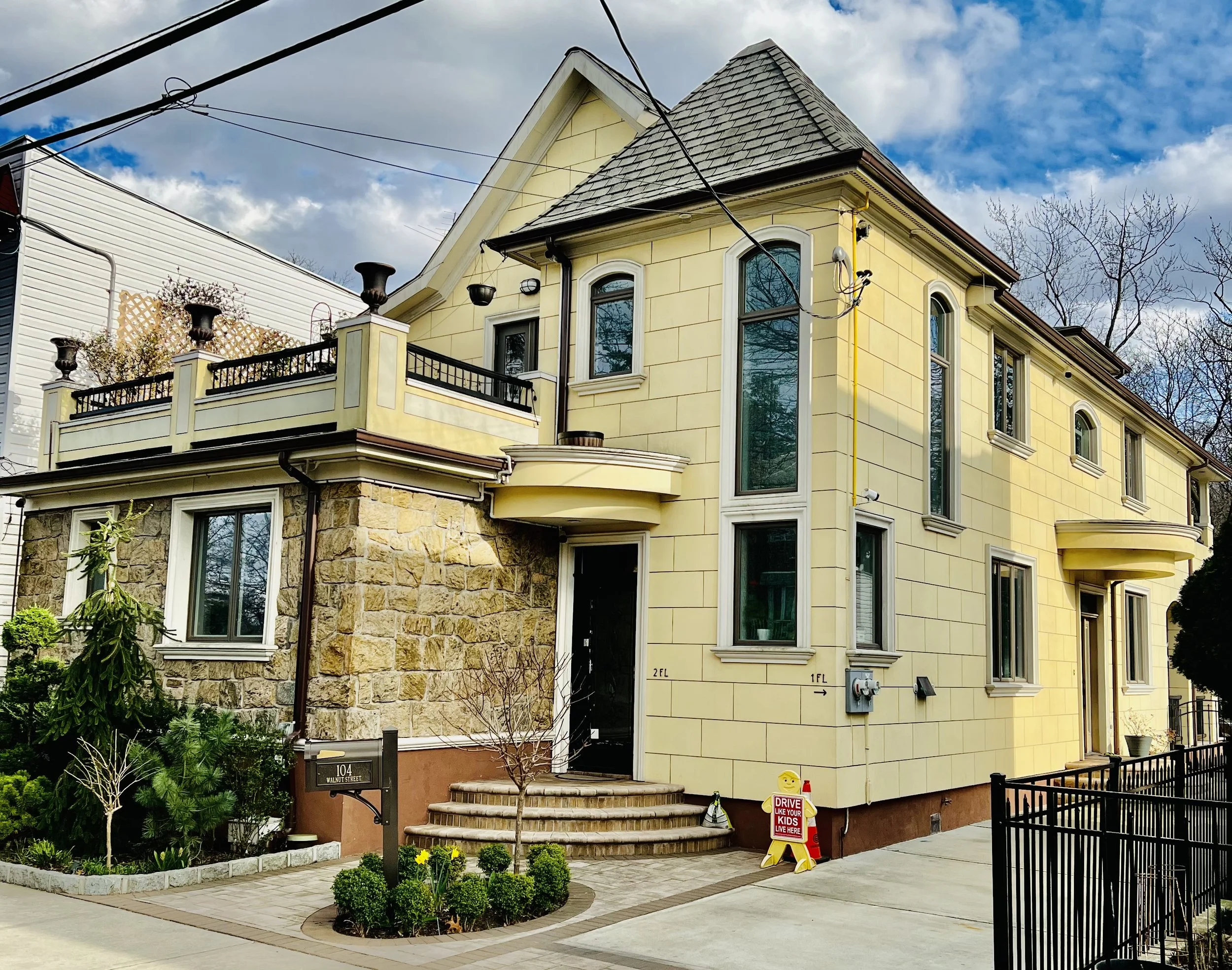 A two-story yellow house with a peaked roof, arched windows, a stone facade on the lower level, and a small front yard with bushes and flowers. There are stairs leading up to the front door and a sign on the sidewalk that reads "Drive Like Your Kids 