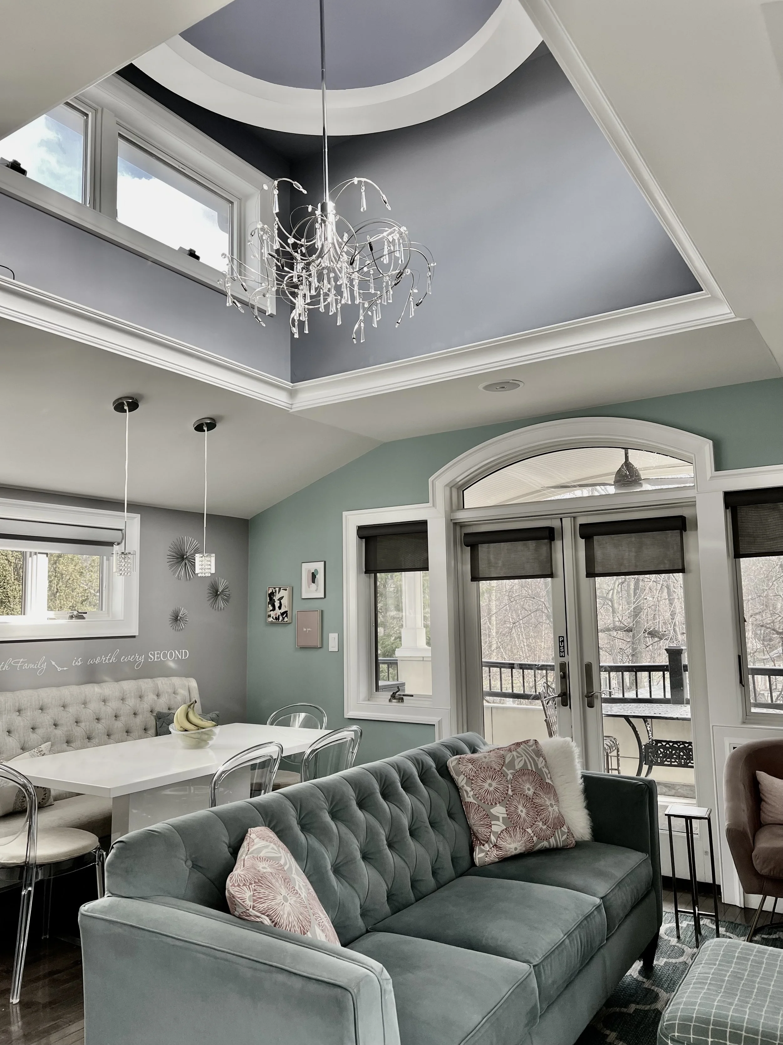 A living room and dining area with a vaulted ceiling, a chandelier, and large windows and doors. There is a gray tufted sofa with pink patterned throw pillows, a white dining table with clear acrylic chairs, and a balcony with outdoor furniture.