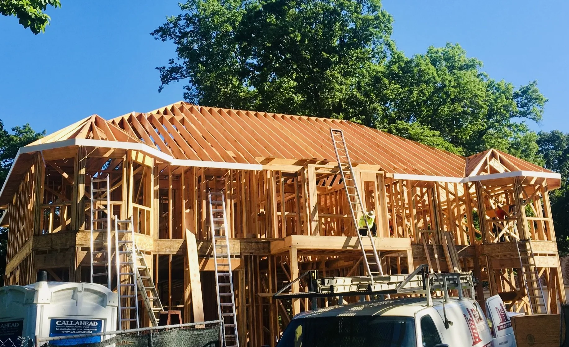 Wooden house under construction with ladders and construction workers, surrounded by green trees and a clear blue sky.
