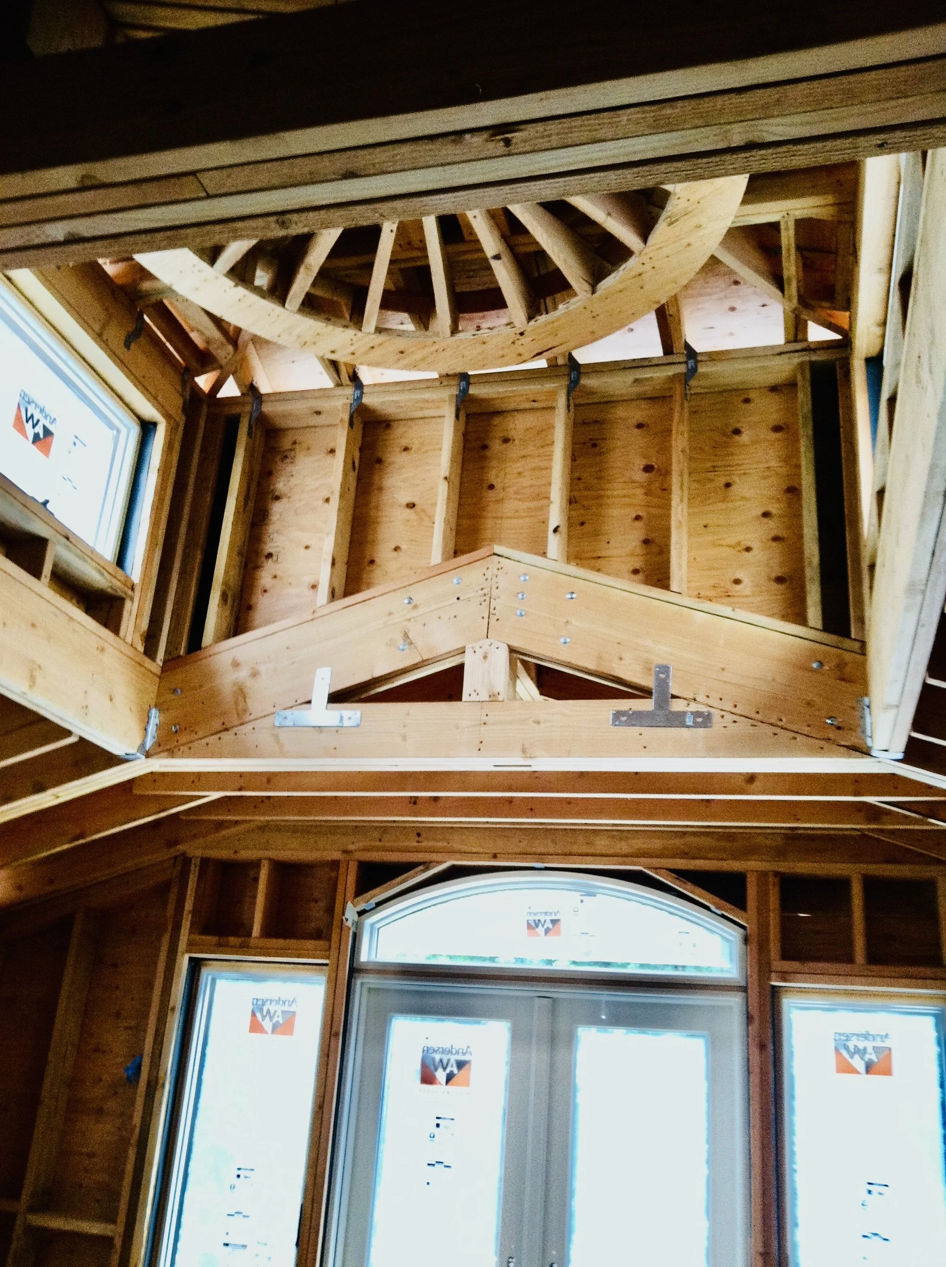 Interior view of a building under construction with exposed wooden framing, large windows, and a curved wood staircase
