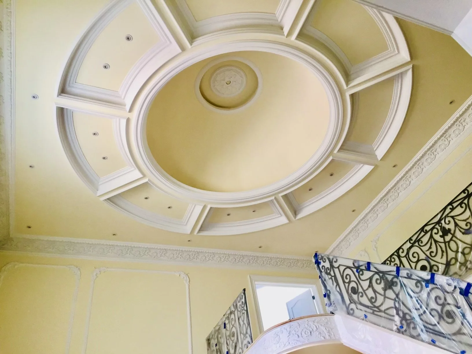 Ceiling with ornate decorative molding and a central recessed circular design, painted in yellow and white, with a part of a staircase with decorative wrought iron railing visible in the lower right corner.