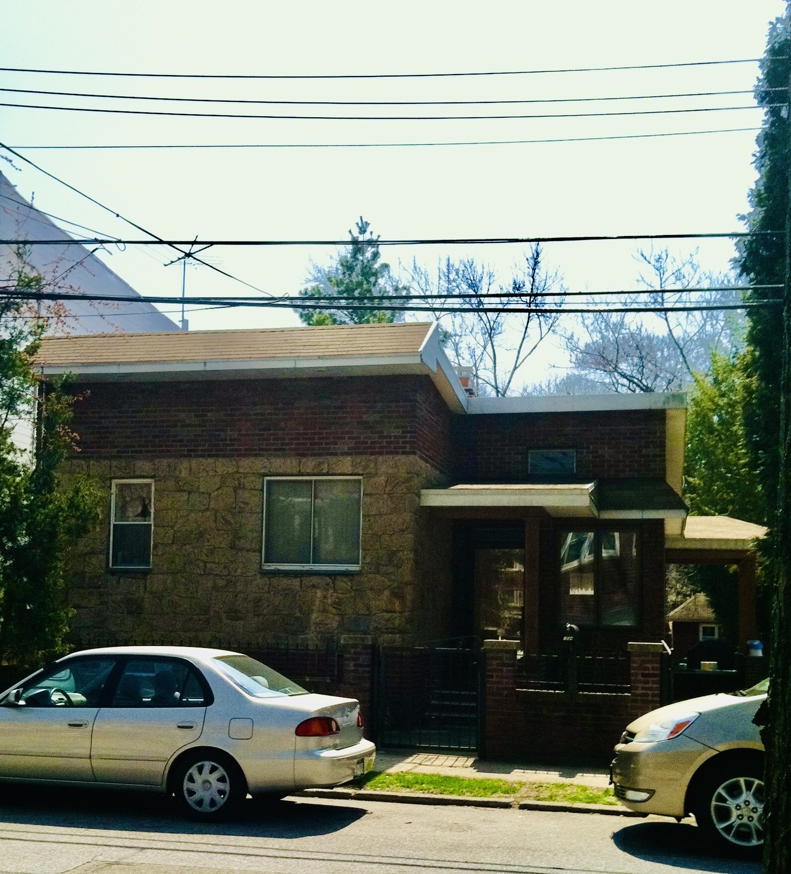 Residential house with a brick facade and stone accents, cars parked in the driveway, trees and power lines in the background, and a clear sky.
