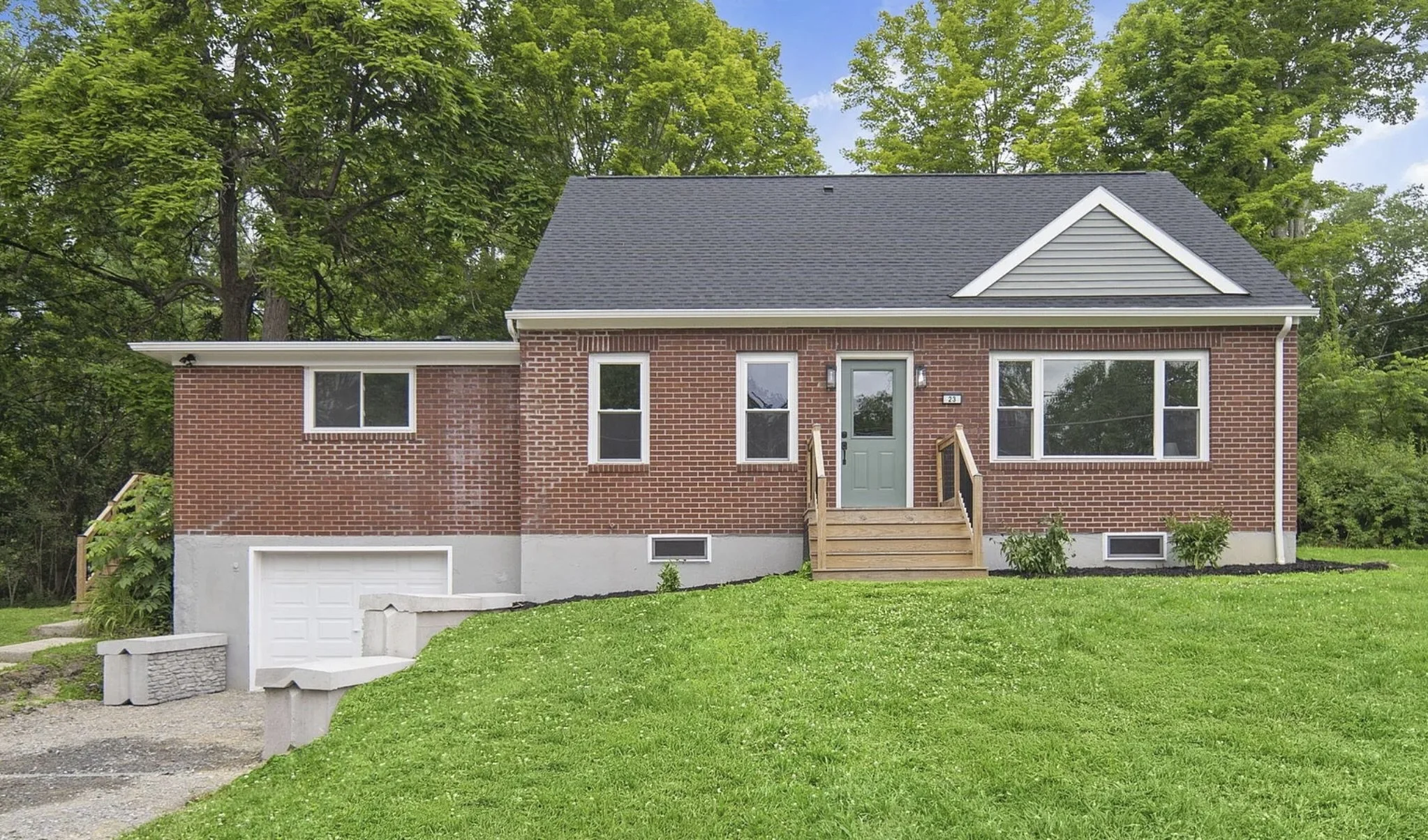 A brick house with a gray roof, multiple windows, a front door with steps, surrounded by green grass and trees in the background.
