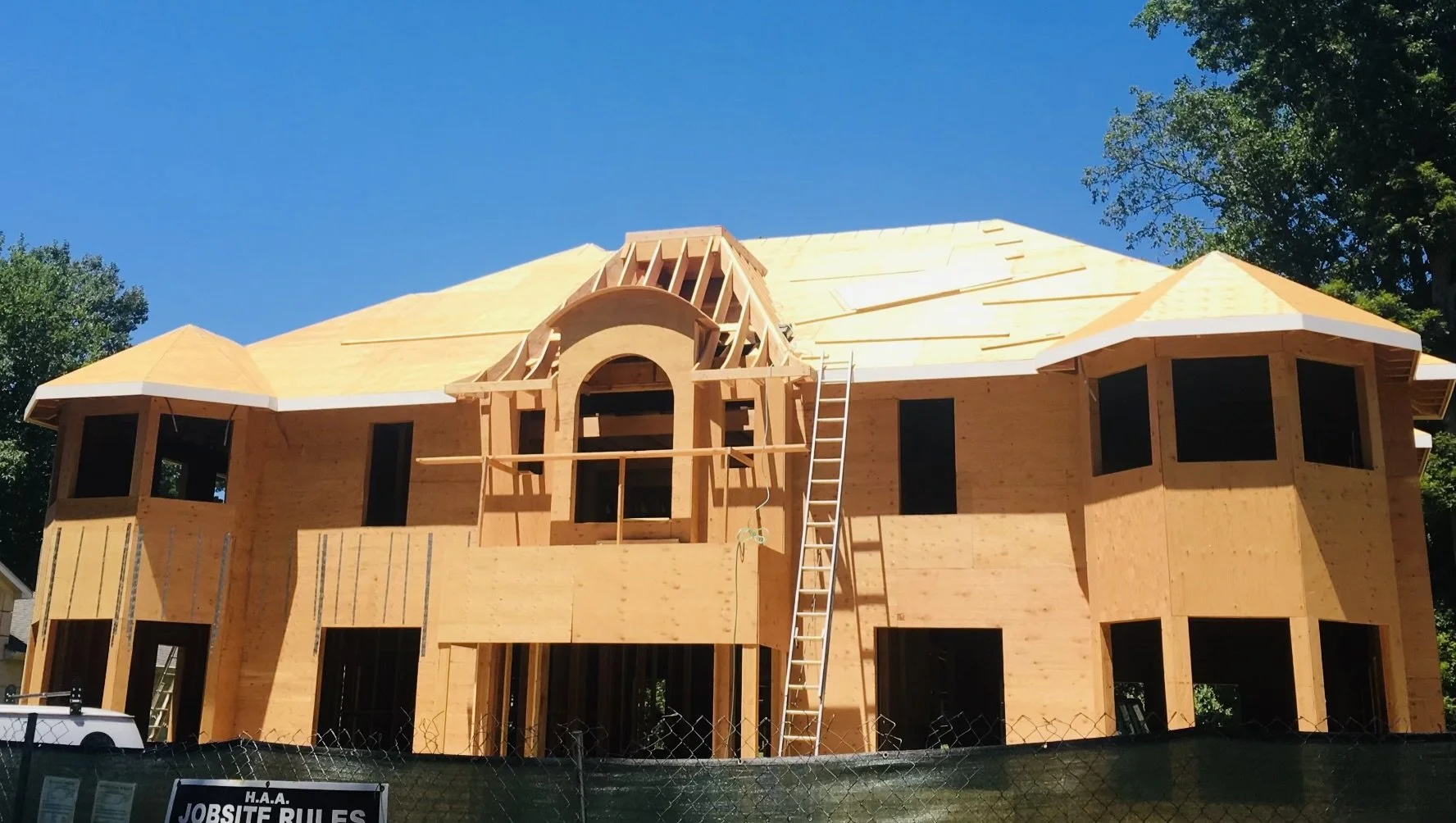 Wooden house under construction with a partially built roof, ladders, and scaffolding in front of a clear blue sky.