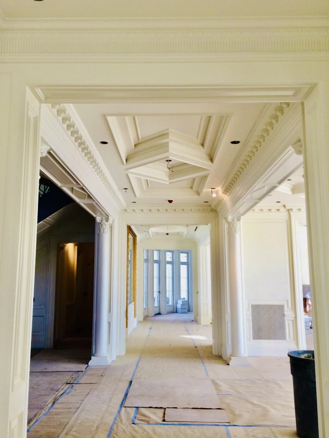 Interior hallway of a luxurious house under renovation, with ornate crown molding, coffered ceiling, tall windows, and protective coverings on the floor.