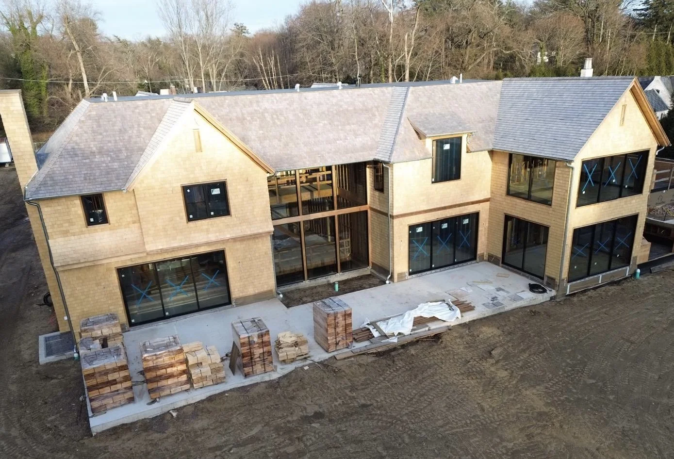 A large house under construction with a brick exterior, multiple large windows, a pitched roof, and a concrete patio in progress. Building materials and stacks of bricks are on the patio.